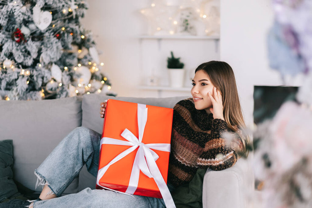 Boldog fehér nő kezében nagy karácsonyi ajándék, miközben ül egy kanapén otthon közel karácsonyfa. Kiváló minőségű fénykép - Fotó, kép