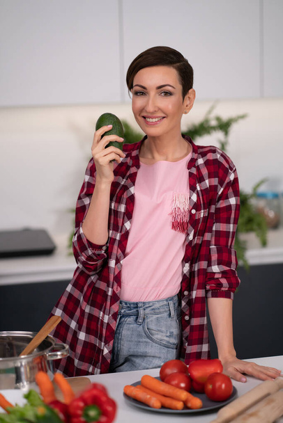 Fresh avocado in the hand of a pretty housewife with a short hairstyle prepares food in the kitchen. Healthy food at home. Healthy food leaving - vegan concept - 写真・画像