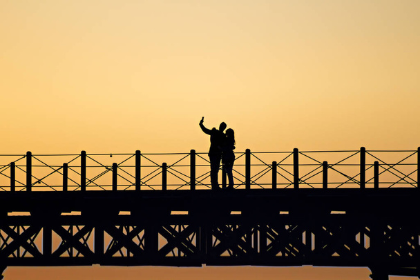 silhouette of a couple taking a selfie in the Tinto Dock "Muelle del Tinto" at sunset in Huelva, Spain - Photo, Image