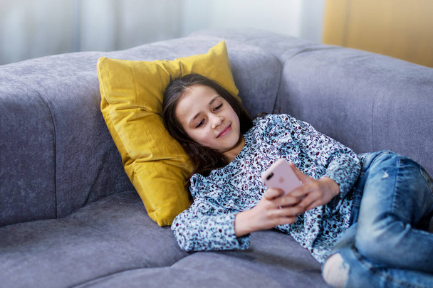 Happy teenage girl checks social networks while holding a smartphone in her hands. A young schoolgirl uses a mobile phone app to play games, correspond with friends, and relax on the couch. - Φωτογραφία, εικόνα