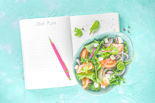 Ένα μπολ από υγιεινή σαλάτα λαχανικών με διάφορα φύλλα σαλάτα, ντομάτες, φέτα, κρεμμύδια, ελαιόλαδο, ανοιχτό μπλε σκυρόδεμα φόντο με σημειωματάριο για Diet Plan αντίγραφο χώρο - Φωτογραφία, εικόνα