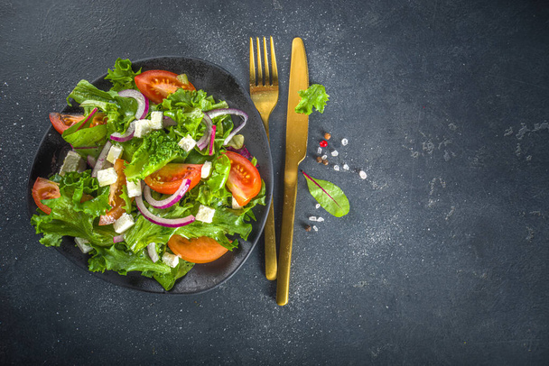 Bowl of healthy vegetable salad with various leaf salad, tomatoes, feta cheese, onions, olive oil, dark grey concrete background copy space - Foto, afbeelding