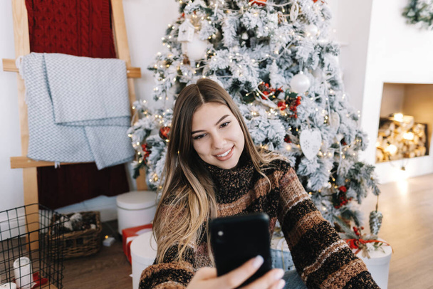 Young caucasian woman wishes merry Christmas by video call using mobile phone in holidays at home. - Foto, afbeelding