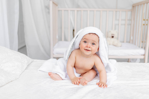 μωρό σε μια πετσέτα στο κρεβάτι σε ένα φωτεινό δωμάτιο κάθεται μετά το μπάνιο - Φωτογραφία, εικόνα