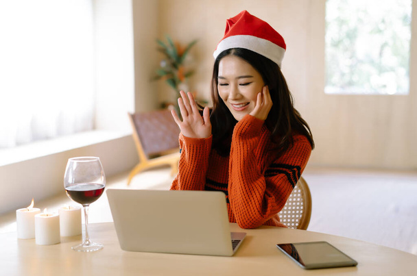 友人や家族とビデオ通話パーティーを作る美しい若いアジアの女性の肖像画は、自宅のリビングルームでクリスマスのお祝いのためのハイウェーブ手を言う。. - 写真・画像