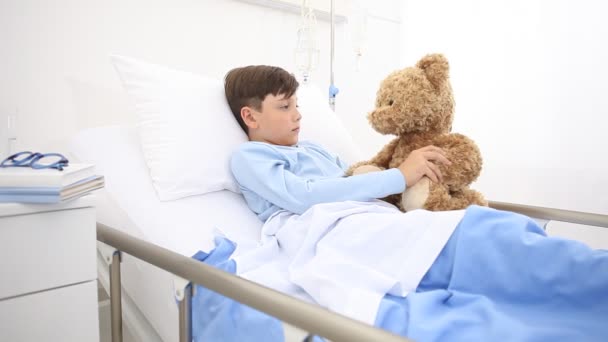 child in hospital lying alone in bed play with teddy bear hugs him and smiling in camera - Footage, Video
