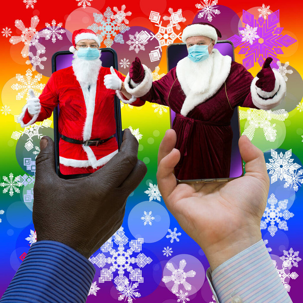 Afroamerican and caucasian-white hands holding phones with 2 Santa Clauses. Santa Clauses have medical masks on. Close-up on a rainbow gradient background with snowflakes. - Photo, Image