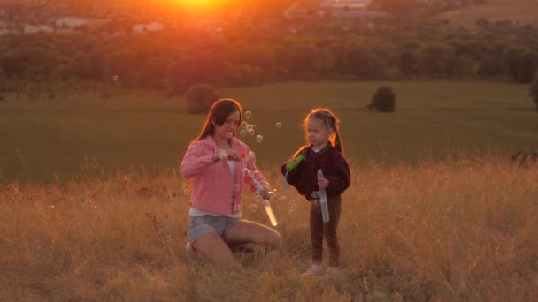 Healthy child and mom are playing. Mother and daughter blow bubbles, baby catches them. Happy family is playing in park at sunset. Family and childhood concept. soap bubbles in sun childrens birthday - Footage, Video