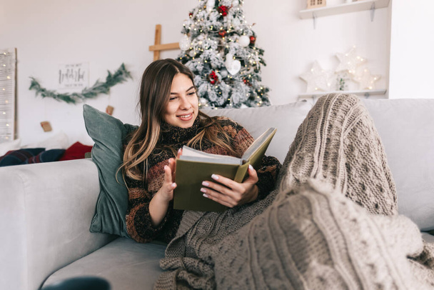 Young caucasian woman relaxing on a sofa at home and reading book in Christmas holidays. High quality photo - Photo, Image