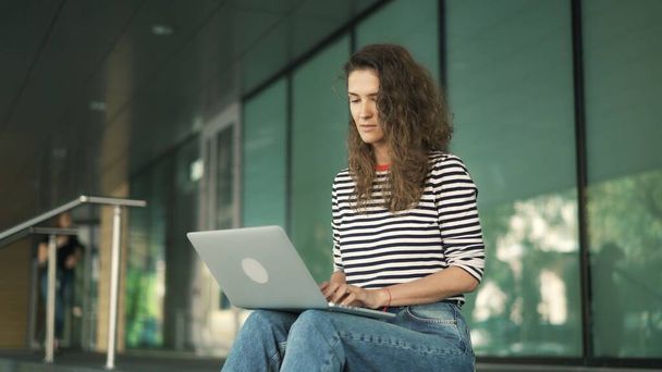 Young concentrated woman curly hair with laptop in stripped sweater, glass office window - Foto, afbeelding