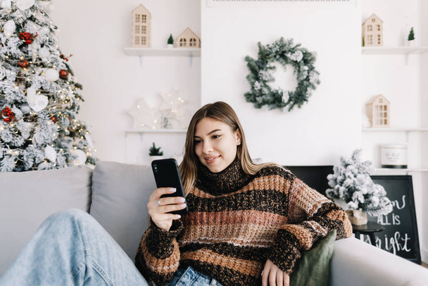 Young smiling caucasian woman using mobile phone in holidays at home on the sofa. High quality photo - Foto, Imagen