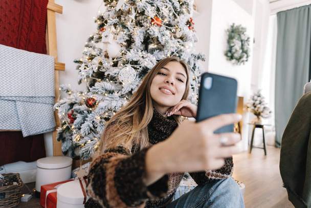 Young smiling caucasian woman making selfie using mobile phone near Christmas tree at home. - Foto, imagen