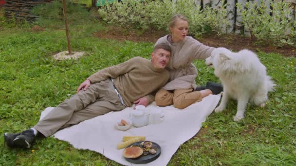 Медленный выстрел. Молодая пара на пикнике, женщина, сидящая на траве, гладящая белую собаку, мужчина, лежащий на женщине. - Кадры, видео