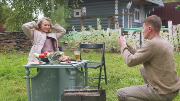 Een vrouw poseert voor haar man op de binnenplaats van de dacha aan een tafel met eten, hij neemt het op een smartphone, slow motion - Video