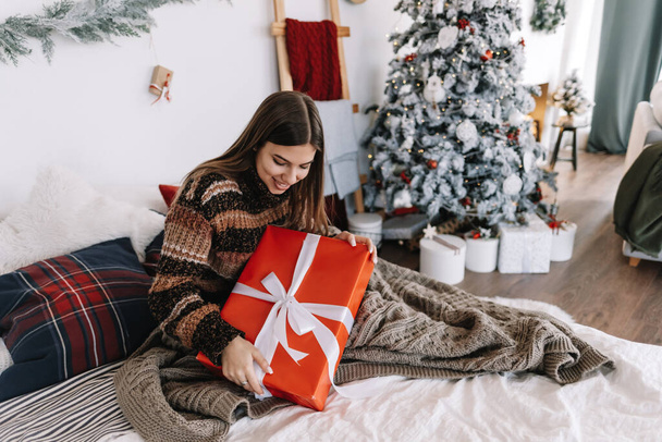 Happy caucasian woman holding big Christmas present while sitting on a bed at home near Christmas tree. - Photo, Image