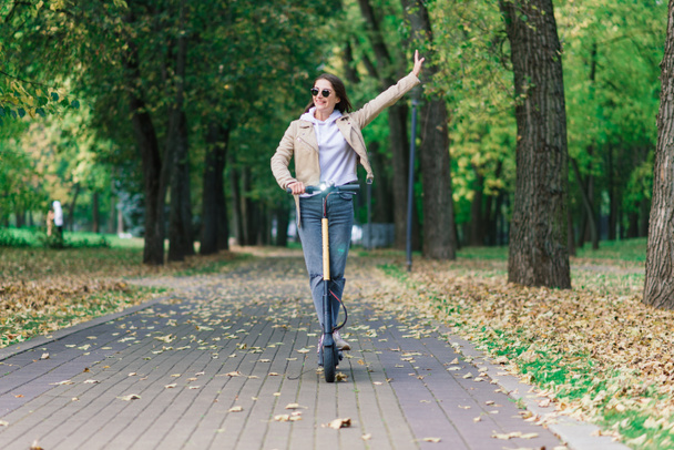 Joven hermosa mujer elegante montar scooter eléctrico en el parque de otoño, transporte ecológico - Foto, imagen
