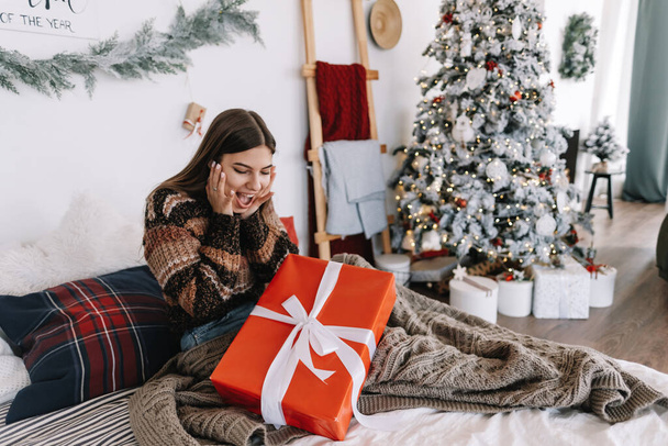 Surprised young caucasian woman with big Christmas present while sitting on a bed at home near Christmas tree. High quality photo - Foto, Bild