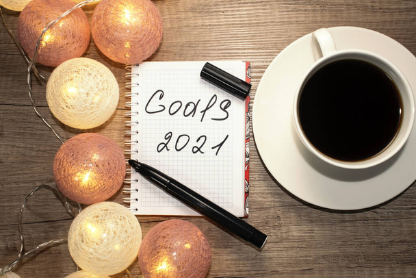 Goals 2021 is written on a Notepad that lies on a wooden table near a white Cup of coffee and Christmas lights. - Foto, Bild