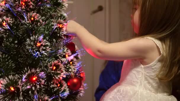 Hijita y madre cuelgan una bola de juguete roja en el árbol de Navidad. Familia feliz, Navidad. Bebé y mamá decoran el árbol de Navidad con bolas de Navidad. Un niño alegre y mamá están jugando junto al árbol. - Metraje, vídeo