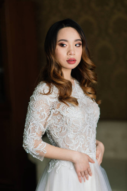 Gorgeous young asian woman in elegant dress looking at camera and window, morning of the bride, wedding - Fotografie, Obrázek