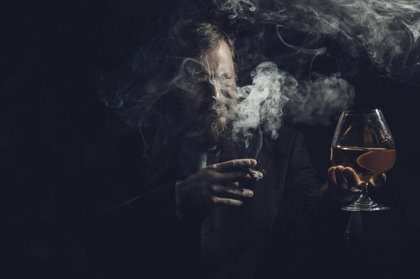Solid confident bearded man in suit with glass of whisky and cigar with fume - Fotografie, Obrázek