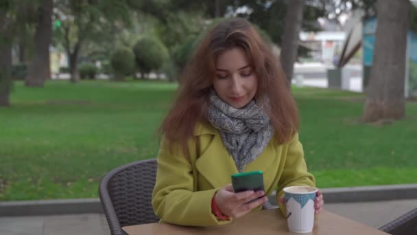 Молодая армянка в кафе на открытом воздухе зимой пьет горячий кофе и общается через видео-чат на смартфоне, улыбается и посылает воздушные поцелуи. - Кадры, видео