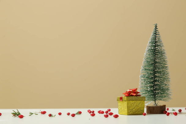 Cute little Christmas tree and gift box with red berries around. Studio shot on beige background. - Photo, Image