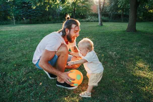 父の日。お父さんは幼児の男の子と屋外でボールをしています。親は公園で子供の息子と一緒に時間を過ごす。本物のライフスタイルの優しい瞬間。幸せなお父さんと活発な家族生活.  - 写真・画像
