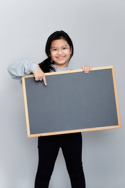 Ένα μικρό κορίτσι κρατά ένα κενό chalkboard θέση για κοινοποίηση. Διάβασε αυτή την ανακοίνωση. Ενημέρωση για το γεγονός. Χαριτωμένη μαθήτρια με μαυροπίνακα. Διαφήμιση. - Φωτογραφία, εικόνα