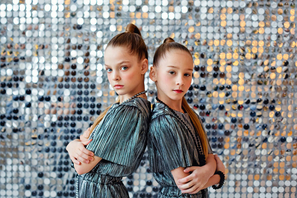 Портрет двух девочек-подростков в блестящих костюмах у мерцающей серебряной стены. Они смотрят в камеру.. - Фото, изображение