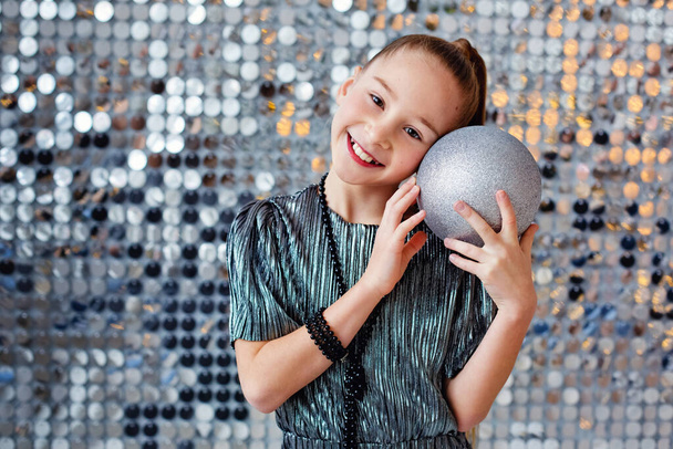 Krásná dívka ve stříbrných šatech s míčem na stříbrném pozadí. Přiloží si balón k obličeji a usměje se.. - Fotografie, Obrázek