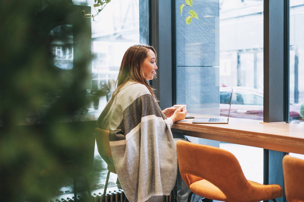 Dorosła uśmiechnięta brunetka w szarym ciepłym szaliku pracująca z laptopem na stole w kawiarni w zimnej porze roku - Zdjęcie, obraz