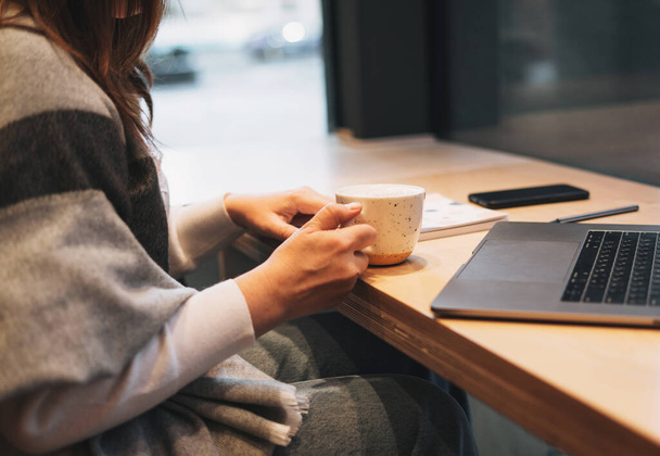 Фото брюнетки в теплом шарфе, работающей с ноутбуком на столе в кафе в холодное время года - Фото, изображение