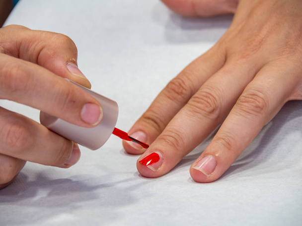 Kobieta malująca paznokcie czerwonym lakierem do paznokci. Zabiegi kosmetyczne salonu piękności. Blisko dziewczyny ręce i paznokcie z pędzlem farby. - Zdjęcie, obraz