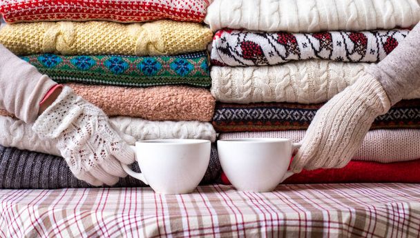 Πολλά πουλόβερ και πουλόβερ Διαφορετικά χρώματα διπλωμένα σε δύο Piles και τα χέρια με δύο φλιτζάνια καφέ. - Φωτογραφία, εικόνα