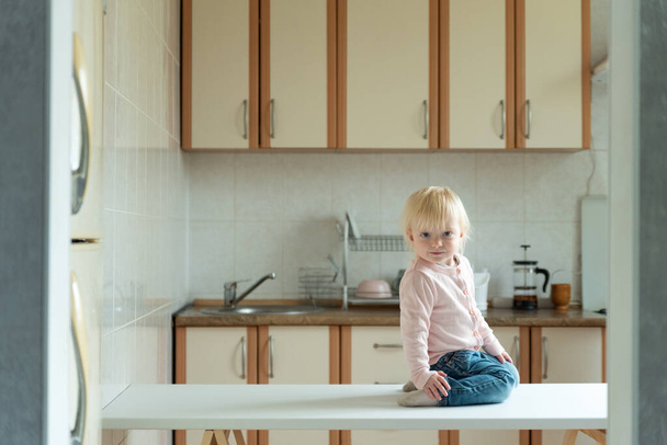 Cute little blonde child sitting on the table in kitchen. Awaiting breakfast. - Photo, Image