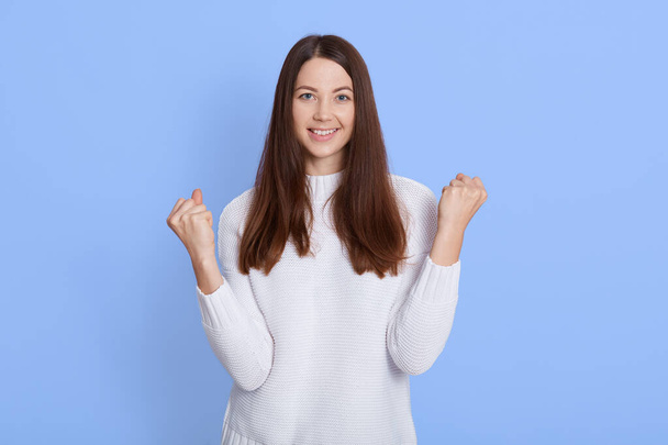 Successful dark haired female, looks happy, clenches fists, accomplishes goal, exclaims finally victory, stands amused over blue background, excited lady dresses white sweater. - Photo, image