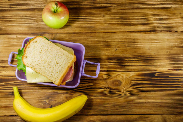 Lunch box with sandwiches, banana and apple on a wooden table. Top view, copy space - Photo, Image
