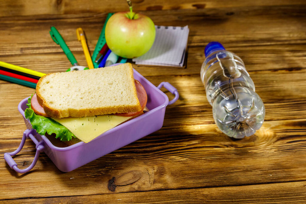 Back to school concept. School supplies, bottle of water, apple and lunch box with sandwiches on a wooden desk - Photo, Image