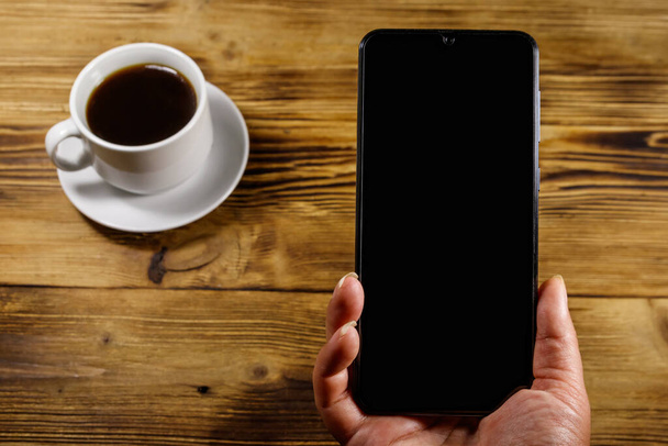 Main avec smartphone moderne et tasse de café sur une table en bois rustique - Photo, image