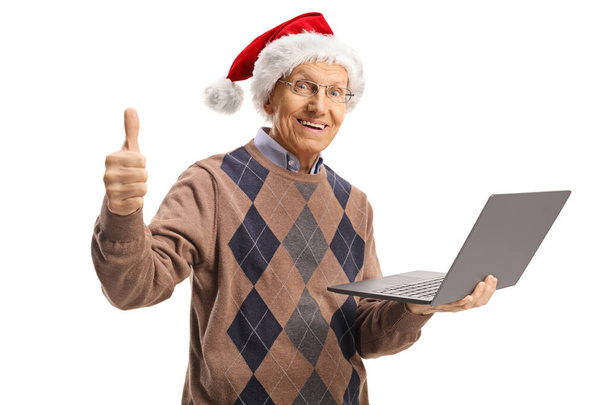 Elderly man with santa claus hat holding a laptop computer and showing thumbs up isolated on white background - Foto, Imagem