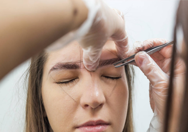 The master corrects the makeup, shapes it with a thread and plucking the eyebrows with tweezers in the beauty salon. Professional face care. - Photo, Image