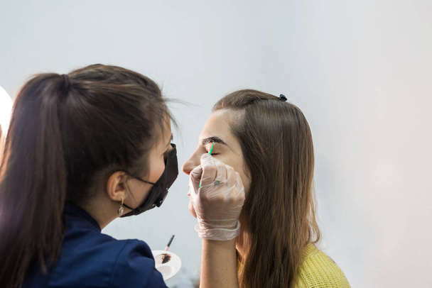 A beautiful young female master makes an eyebrow correction procedure to a client. Eyebrow dyeing concept in beauty salon. - Foto, Imagem