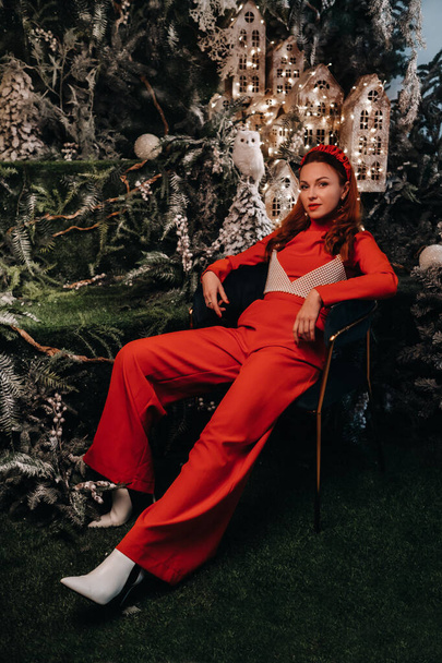 Kırmızı elbiseli bir kız Noel arka planında bir sandalyede oturuyor. Noel ağaçlarının ve küçük Noel evlerinin arka planında kırmızılı bir kadın.. - Fotoğraf, Görsel