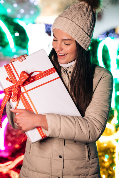 Happy young woman in hat holding christmas present box while standing on street. Winter concept. - Fotoğraf, Görsel
