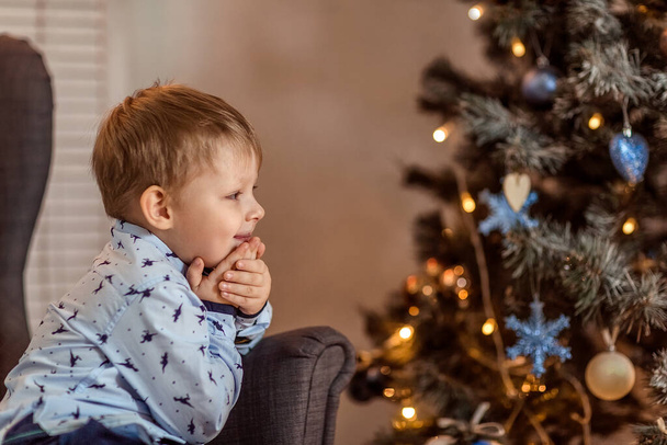 Un hermoso niño de 4-5 años de edad se encuentra (se sienta) cerca del sofá y sueña con unas vacaciones y regalos. El concepto de vacaciones de invierno. Enfoque suave selectivo, bokeh (fondo borroso). - Foto, Imagen