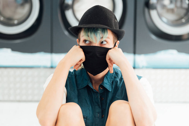 Young pretty Caucasian woman with a casual look who is bored, fatigued in a public laundry. She wears a face mask to protect herself from the coronavirus. - Photo, Image