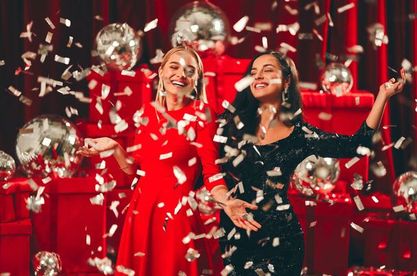 Bonne année à vous ! Deux belles femmes sexy dansant en confettis. Fête du Nouvel An. La veille de Noël. Sur fond de boîtes-cadeaux rouges et de boules de disco. - Photo, image
