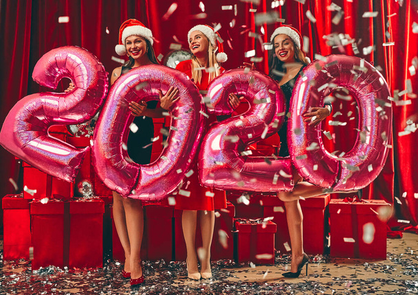 Frohes neues Jahr! Drei schöne sexy Frauen in Nikolausmützen neben roten Geschenkboxen mit aufgeblasenen Luftballons mit der Nummer 2020. Neujahrsparty. Heiligabend. - Foto, Bild