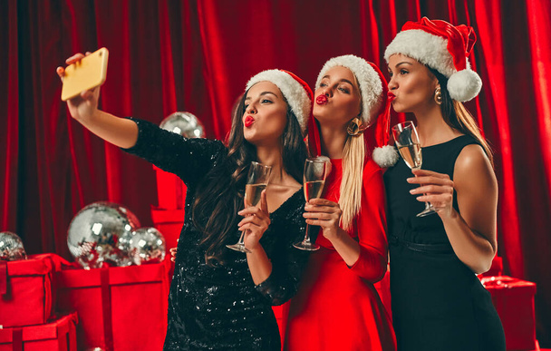 Bonne année à vous ! Trois belles femmes sexy en chapeaux de Père Noël avec des verres de champagne prennent un selfie. Fête du Nouvel An. La veille de Noël. Sur fond de boîtes-cadeaux rouges et de boules de disco. - Photo, image